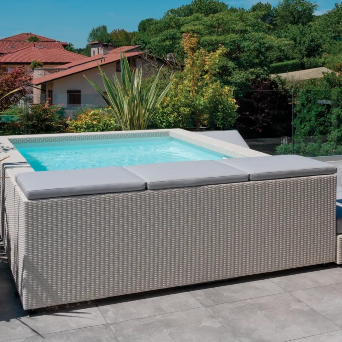 Cuscino bordo minipiscina Laghetto Playa Una minipiscina Laghetto Playa piscina fuori terra di design, integrata in un'area esterna con pavimentazione in piastrelle grigie. La piscina è caratterizzata da un rivestimento esterno in fibra sintetica intrecciata a mano colore binco, con una texture intrecciata. La piscina ha una forma rettangolare con angoli arrotondati. L'acqua è di un colore azzurro chiaro e sembra pulita e invitante. Il bordo della piscina è realizzato sempre in fibra sintetica intrecciata continuativo del rivestimento esterno. Sulla parte superiore del rivestimento, è presente un cuscino lungo rettangolare di colore grigio chiaro. Il cuscino è realizzato in materiale Sky nautico adatto all'esterno. La piscina è circondata da una pavimentazione esterna in piastrelle grigie di grande formato. Lo sfondo mostra un'area verde con alberi e arbusti, creando un'atmosfera naturale e rilassante. Si intravedono anche i tetti di alcune case in lontananza.