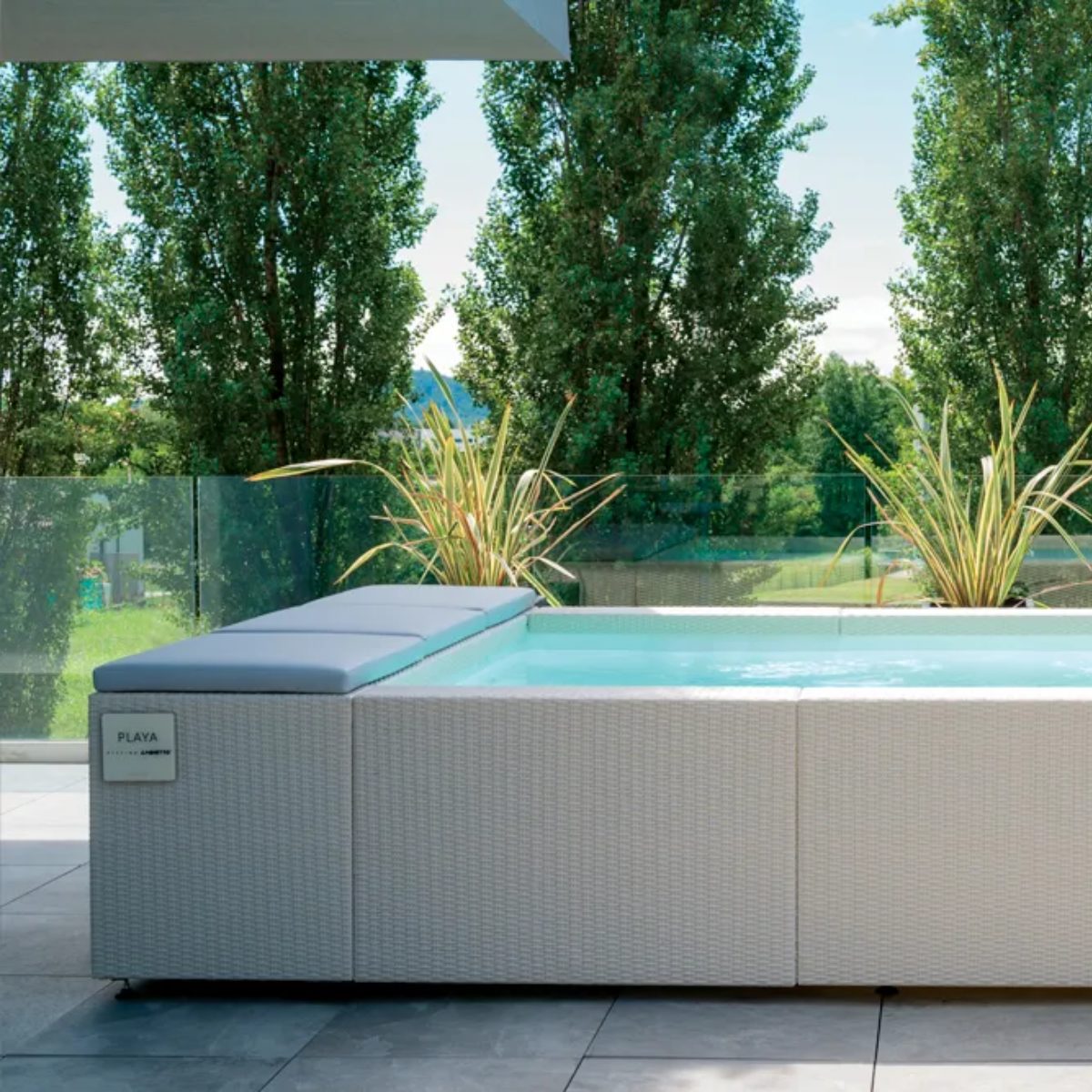 Piscina Laghetto Playa bianca modello due con cuscino prendisole azzurro Una piscina fuori terra con idromassaggio integrato, modello Laghetto Playa 2 (220 x 320 cm), è posizionata su una pavimentazione in piastrelle grigie. La piscina ha un rivestimento esterno in vimini sintetico chiaro e cuscini di seduta coordinati. Sullo sfondo, alberi alti e rigogliosi creano un'atmosfera rilassante. L'immagine mostra il logo "PLAYA" su un lato della piscina.