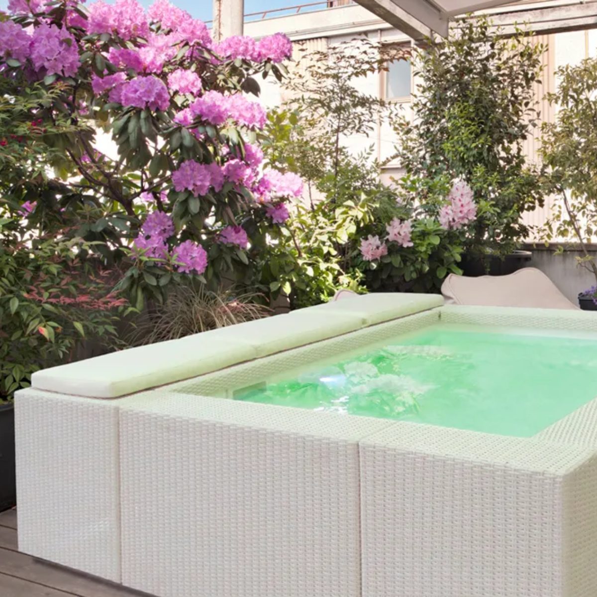 Piscina Laghetto Playa bianca modello uno con cuscino prendisole bianco Una piscina Laghetto Playa modello 1 (220x220 cm) quadrata fuori terra, con rivestimento in vimini chiaro e cuscino di seduta, è circondata da una rigogliosa vegetazione fiorita. L'acqua della piscina è verde chiaro e sembra invitante. La scena si svolge su una terrazza o un balcone, con un'atmosfera rilassante e naturale.