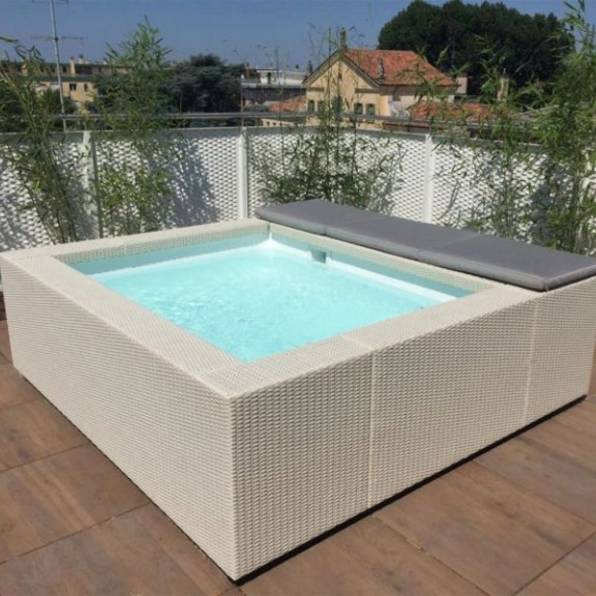 Piscina Laghetto Playa bianca modello uno con cuscino prendisole grigio Una piscina fuori terra modello Laghetto Playa modello Playa 1 (220x220 cm) di forma quadrata con idromassaggio integrato è posizionata su una terrazza con pavimentazione in piastrelle. La piscina ha un rivestimento esterno in vimini sintetico chiaro e un cuscino di seduta grigio su un lato. L'acqua è di un colore azzurro chiaro. Sullo sfondo, si vedono edifici residenziali e vegetazione.