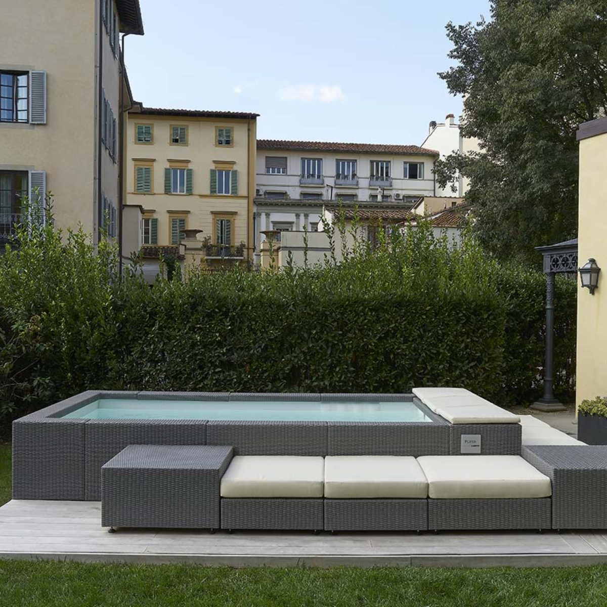 Piscina Laghetto Playa grigio con cuscino prendisole bianco e living La piscina laghetto Playa è una struttura fuori terra di forma rettangolare con angoli leggermente arrotondati, integrata in un'area esterna con un design moderno e curato. La piscina laghetto Playa modello 3 è di forma rettangolare compatta, ideale per spazi esterni limitati, in questa foto è abbellita dal Living esterno e cuscini. Il rivestimento esterno, il bordo e gli accessori del living sono realizzato in fibra sintetica intrecciata di colore grigio, simile al vimini sintetico, che conferisce un aspetto elegante e contemporaneo. L'acqua della piscina è di un colore azzurro chiaro, che suggerisce pulizia e freschezza, colore dato dal rivestimento interno in PVC bianco La piscina è integrata in un'area esterna con pavimentazione in piastrelle chiare e un'area relax adiacente, composta da sedute imbottite nello stesso materiale intrecciato del rivestimento della piscina. La piscina è situata in un giardino con vegetazione rigogliosa, che crea un'atmosfera rilassante e privata. Sullo sfondo, si vedono edifici residenziali che suggeriscono un contesto urbano o semi-urbano. La piscina ha un aspetto moderno, elegante e funzionale. L'integrazione con l'area relax e l'ambiente circostante crea uno spazio esterno armonioso e accogliente.