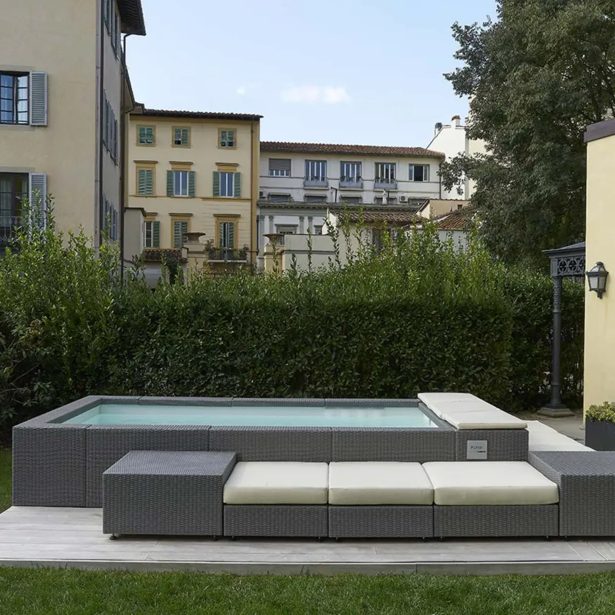 Piscina Laghetto Playa grigio con cuscino prendisole bianco e living La piscina laghetto Playa è una struttura fuori terra di forma rettangolare con angoli leggermente arrotondati, integrata in un'area esterna con un design moderno e curato. La piscina laghetto Playa modello 3 è di forma rettangolare compatta, ideale per spazi esterni limitati, in questa foto è abbellita dal Living esterno e cuscini. Il rivestimento esterno, il bordo e gli accessori del living sono realizzato in fibra sintetica intrecciata di colore grigio, simile al vimini sintetico, che conferisce un aspetto elegante e contemporaneo. L'acqua della piscina è di un colore azzurro chiaro, che suggerisce pulizia e freschezza, colore dato dal rivestimento interno in PVC bianco La piscina è integrata in un'area esterna con pavimentazione in piastrelle chiare e un'area relax adiacente, composta da sedute imbottite nello stesso materiale intrecciato del rivestimento della piscina. La piscina è situata in un giardino con vegetazione rigogliosa, che crea un'atmosfera rilassante e privata. Sullo sfondo, si vedono edifici residenziali che suggeriscono un contesto urbano o semi-urbano. La piscina ha un aspetto moderno, elegante e funzionale. L'integrazione con l'area relax e l'ambiente circostante crea uno spazio esterno armonioso e accogliente.