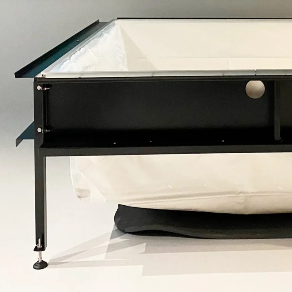 Struttura minipiscina Laghetto Playa Un angolo di una struttura rettangolare della vasca Playa. La struttura è caratterizzata da una cornice metallica nera, con un profilo a forma di L che definisce l'angolo, e da un rivestimento interno bianco, realizzato in tessuto Trevira spalmato da 850 g/mq. Si notano viti e bulloni che tengono insieme i vari elementi della struttura metallica, un ripiano orizzontale sotto il rivestimento interno e un piedino regolabile con una base rotonda in gomma. L'immagine, molto dettagliata, mostra la struttura in modo chiaro e preciso, con una composizione semplice e minimalista che focalizza l'attenzione sull'angolo.