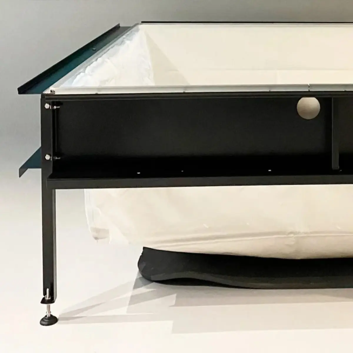 Struttura minipiscina Laghetto Playa Un angolo di una struttura rettangolare della vasca Playa. La struttura è caratterizzata da una cornice metallica nera, con un profilo a forma di L che definisce l'angolo, e da un rivestimento interno bianco, realizzato in tessuto Trevira spalmato da 850 g/mq. Si notano viti e bulloni che tengono insieme i vari elementi della struttura metallica, un ripiano orizzontale sotto il rivestimento interno e un piedino regolabile con una base rotonda in gomma. L'immagine, molto dettagliata, mostra la struttura in modo chiaro e preciso, con una composizione semplice e minimalista che focalizza l'attenzione sull'angolo.