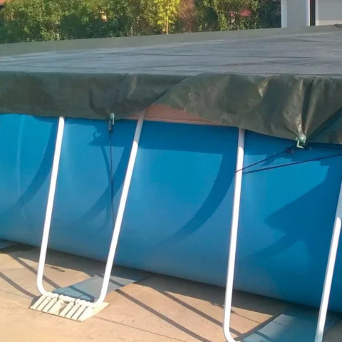 Piscina fuori terra di colore blu, presenta una struttura di supporto esterna composta da tubi metallici bianchi verticali e orizzontali. La piscina è coperta da un telo di copertura invernale di colore verde scuro. Il telo è steso sopra la superficie dell'acqua e si estende oltre i bordi della piscina. È fissato alla struttura della piscina tramite un sistema di elastici neri con ganci in plastica verde. Si possono notare diversi di questi elastici che collegano gli occhielli presenti sul bordo del telo ai montanti verticali della piscina. La piscina è posizionata su una superficie pavimentata esterna, probabilmente un patio o un terrazzo, di colore chiaro. Sullo sfondo si possono notare alberi e vegetazione, suggerendo un ambiente esterno residenziale.