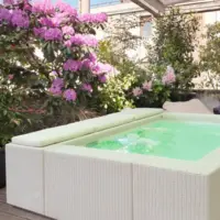 Piscina fuori terra Laghetto Playa con rivestimento in fibra sintetica intrecciata effetto rattan. Presentata in un contesto outdoor. Colore rivestimento bianco, colore cuscino bianco.