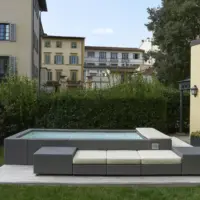 Piscina fuori terra Laghetto Playa con rivestimento in fibra sintetica intrecciata effetto rattan. Presentata in un contesto outdoor. Colore rivestimento grigio, colore cuscino bianco.