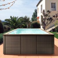 Piscina fuori terra Laghetto Dolcevita Rattan con rivestimento in fibra sintetica intrecciata effetto rattan e bordo in ecolegno. Piscina posizionata in giardino di casa privata. Colore rivestimento esterno e bordo in ecolegno marrone.