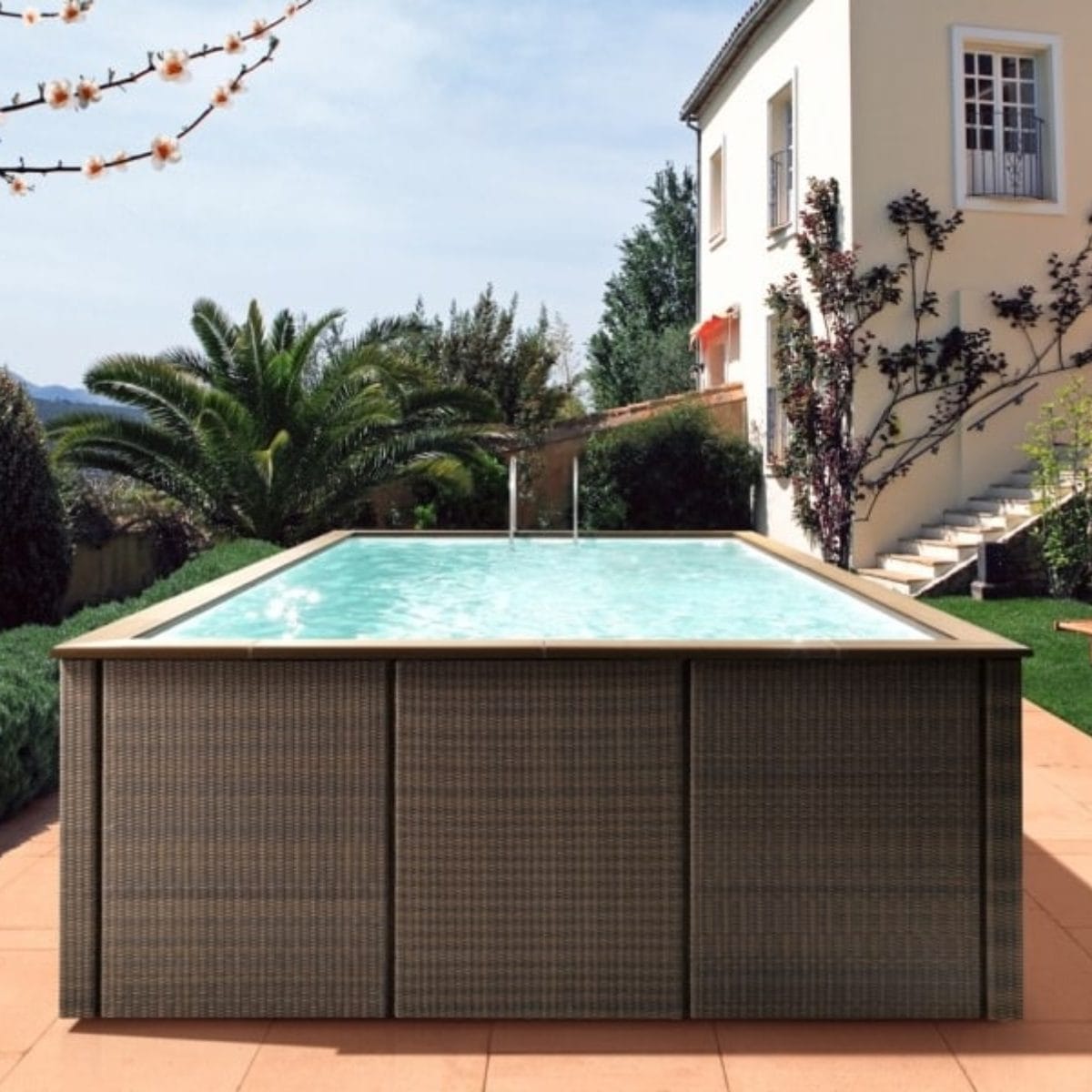 Piscina fuori terra Laghetto Dolcevita Rattan con rivestimento in fibra sintetica intrecciata effetto rattan e bordo in ecolegno. Piscina posizionata in giardino di casa privata. Colore rivestimento esterno e bordo in ecolegno marrone.