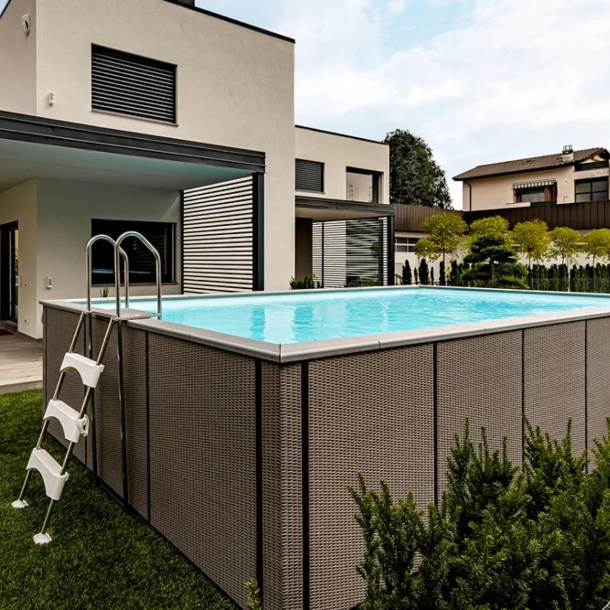 Piscina fuori terra Laghetto Dolcevita Rattan con rivestimento in fibra sintetica intrecciata effetto rattan e bordo in ecolegno. Piscina posizionata in giardino di casa privata. Colore rivestimento esterno e bordo in ecolegno grigio.